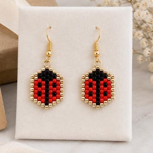 May include: A pair of dangle earrings featuring a hexagonal ladybug design crafted from small red, black, and gold beads. The earrings hang from gold-toned metal hooks and are displayed on a white card next to a brown gift tag with the text LittleSparkArtStudio.