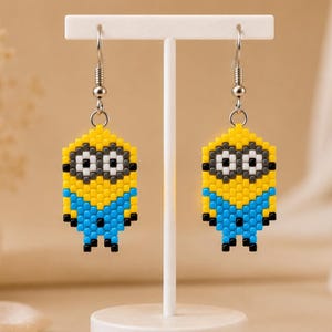 Peut inclure: Une paire de boucles d'oreilles pendantes fabriquées à partir de petites perles carrées dans un style pixelisé. Le motif représente un personnage jaune, bleu, noir, blanc et gris avec de grandes lunettes circulaires, suspendu à des crochets en métal argenté.