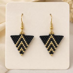 Può includere: Un paio di orecchini a forma di triangolo realizzati con piccole perline nere e oro metallizzato. Gli orecchini presentano un motivo geometrico a chevron e sono appesi a ganci in metallo dorato, fissati su un cartoncino quadrato.
