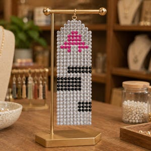 May include: A rectangular beaded tapestry hanging from a gold-toned metal T-bar stand. The piece is constructed from clear, translucent beads with a pattern of hot pink beads at the top and black beads arranged in geometric blocks throughout the center.