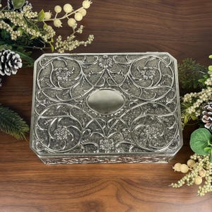 May include: A rectangular silver-toned metal box featuring an intricate embossed pattern of swirling vines and flowers across the lid. The center of the lid contains a smooth, plain oval plaque. The box has a textured background finish.