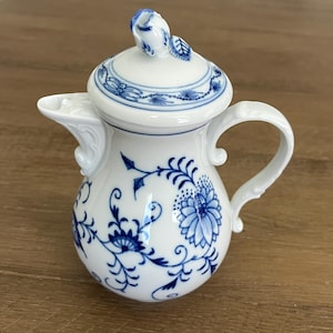 Puede incluir: Una jarra de crema de porcelana blanca con un patrón floral azul, un asa curva y una tapa rematada con un pequeño capullo de rosa esculpido. El cuerpo está decorado con intrincados motivos botánicos azules y enredaderas.