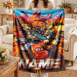 May include: A fleece blanket featuring a colourful graphic print of various animated race cars from the movie Cars, set against a desert landscape with rock formations. The word NAME is printed in large, bold, metallic-style letters at the bottom.