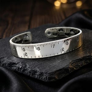 May include: A silver cuff bracelet featuring a polished finish and a design resembling a ruler with engraved black markings and numbers from 2 to 9. The interior of the band displays the hallmark S925, indicating the metal composition.