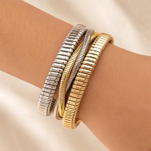 Puede incluir: Un conjunto de tres pulseras de metal flexible con un diseño apilable. Una pulsera es de tono plateado con eslabones rectangulares, otra es una cadena de serpiente plateada fina y otra es de tono dorado con eslabones rectangulares, todas en la muñeca.