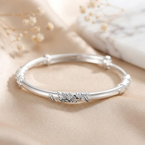 Pode incluir: Um bracelete de prata polida com uma banda lisa e arredondada, apresentando segmentos decorativos integrados. Estes segmentos exibem entalhes detalhados em relevo de pequenas flores e folhas, criando um contraste texturizado contra a superfície brilhante e reflexiva do metal.