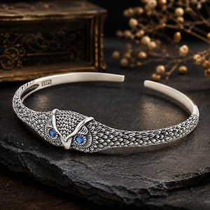 May include: A silver cuff bracelet featuring a detailed owl head centerpiece with two small blue gemstone eyes. The band is textured with a feather-like pattern and includes an S925 hallmark stamp on the interior surface of the metal.
