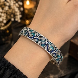 Pode incluir: Uma pulseira tipo bracelete de prata com um intrincado padrão de filigrana vazada, preenchido com espirais de esmalte azul e turquesa vibrantes. O metal apresenta um acabamento texturizado e oxidado dentro dos elementos de design curvos e entrelaçados.