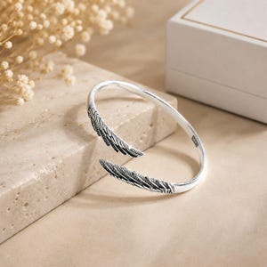 Puede incluir: Una pulsera abierta de tono plateado con dos extremos detallados en forma de ala. La superficie metálica muestra intrincados grabados de plumas con acentos oscuros a lo largo de cada ala. Un pequeño sello rectangular con 925 es visible.