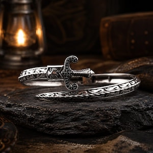 May include: A silver-toned metal cuff bracelet featuring a sword-shaped centerpiece with black stone accents. The band displays an intricate engraved pattern along its length, and the central sword motif includes a small eagle emblem detail.