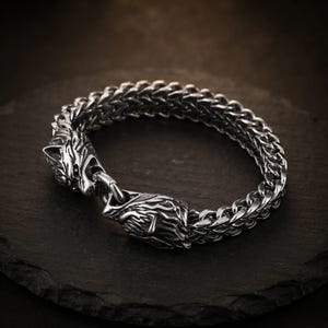 May include: A silver-coloured metal chain bracelet featuring a thick, woven wheat-link design. The two ends of the bracelet terminate in detailed, sculpted wolf head motifs that meet at the centre, serving as the primary focal point of the piece.