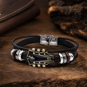 Op de afbeelding: Een meerstrengs zwarte leren armband met een centrale zwart-gouden gitaarkop-bedel voorzien van een vioolsleutel. Het ontwerp bevat zilverkleurige metalen kralen, donkerbruine houten ringen en een roestvrijstalen sluiting.