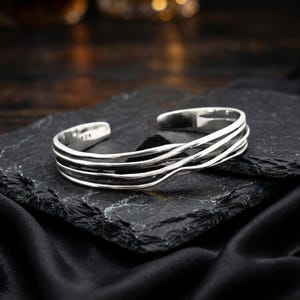 May include: A silver cuff bracelet featuring four thin, smooth, parallel bands that curve and overlap slightly. The metal has a polished, reflective finish, and the open-ended design allows for adjustment. The bracelet is a solid, continuous piece.