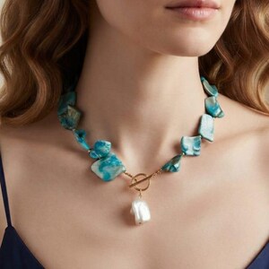 May include: A necklace featuring irregular, marbled turquoise-blue stone beads strung together. The piece includes a gold-toned metal toggle clasp at the center, from which a single, large, white, organic-shaped baroque pearl pendant hangs.