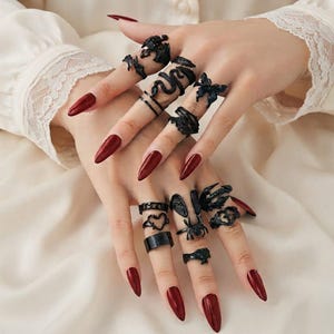 May include: A collection of black metal rings featuring various designs including a snake, a butterfly, a spider, a heart, wings, and textured bands. The rings are worn across multiple fingers, showcasing intricate details and dark finishes.