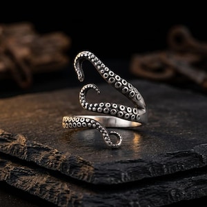 Op de afbeelding: Een zilverkleurige ring in de vorm van een inktvistentakel die om de vinger krult. Het metalen oppervlak heeft een donkere, geoxideerde afwerking in de verdiepte delen en verhoogde, cirkelvormige zuignappen langs het tentakelontwerp.