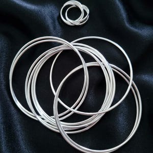May include: A set of seven interlocking silver-toned bangles arranged in a circular cluster, accompanied by a smaller, separate ring composed of three thin, intertwined silver-toned bands, both featuring a smooth, polished metallic finish.