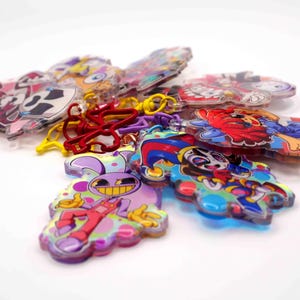 Könnte beinhalten: Eine Sammlung von Acryl-Schlüsselanhängern mit farbenfrohen Cartoon-Charakter-Illustrationen und lebendigen Designs. Jedes Stück enthält einen Metallverschluss in verschiedenen Farben wie Rot, Gelb und Lila, der an transparenten, gestanzten Acrylformen befestigt ist.