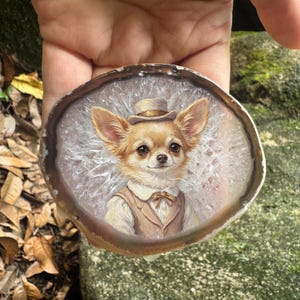 Puede incluir: Un retrato pintado a mano de un chihuahua de color canela con chaleco, camisa blanca de cuello, pajarita y sombrero de copa, sobre una rodaja circular de ágata translúcida y cristalina con un borde marrón oscuro y un centro blanco.