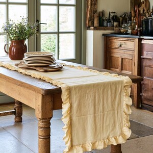 Puede incluir: Un camino de mesa rectangular de color amarillo pálido con un borde de tela con volantes. El camino está colocado sobre una mesa de madera, debajo de una pila de platos de cerámica y una jarra de cerámica marrón con flores azules.