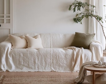 May include: A three-seater sofa covered in a cream-colored linen slipcover, featuring two matching cream throw pillows and one olive green square throw pillow. The sofa has a relaxed, draped fit with visible seams and a soft, textured fabric.