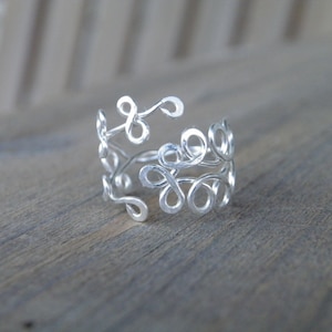 May include: A silver ring with a delicate, intricate design of interwoven loops and swirls. The ring is made of thin wire and has a delicate, feminine look.