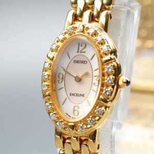 May include: A gold-tone Seiko Exceline wristwatch featuring an oval-shaped case surrounded by a bezel set with small clear stones. The watch has a cream-coloured dial with gold-tone hands and numerals at the 12, 3, 6, and 9 o'clock positions.