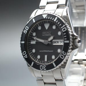 Puede incluir: Un reloj automático Seiko de acero inoxidable con esfera negra y bisel giratorio negro. La esfera presenta marcadores de hora luminosos, manecillas plateadas y una ventana de día y fecha en la posición de las tres. Es water 10 bar resist.
