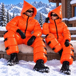 Op de afbeelding: Twee personen in feloranje, glanzende, gewatteerde jumpsuits met puntige kappen, zwarte ritsen, zwarte handschoenen en zwarte laarzen. De jumpsuits zijn voorzien van meerdere zwarte gespriemen langs de benen en hebben een opvallende glans.