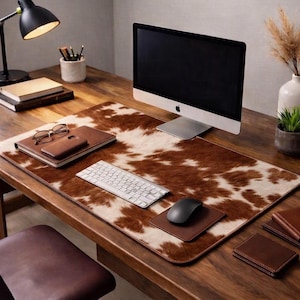 Peut inclure: Un grand tapis de bureau rectangulaire avec un motif de peau de vache marron et blanc. Le tapis est posé sur un bureau en bois et supporte un clavier sans fil argenté, une souris d'ordinateur gris foncé sur un tapis de souris marron.