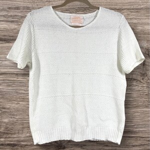 May include: A short-sleeved white knit top with a round neckline and a textured, open-weave pattern. The garment features a ribbed hem and a small rectangular brand label sewn inside the collar that reads Country Sophisticates.