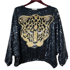May include: A black long-sleeve top covered in black sequins, featuring a large, centered leopard head graphic made of gold and black sequins. The garment has a wide, loose fit with a rounded neckline and is displayed on a dark brown hanger.