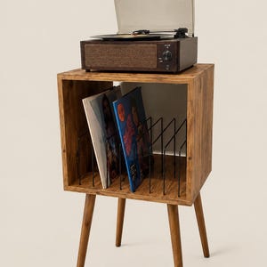 May include: A wooden record player stand with four tapered legs and an open storage compartment featuring black metal dividers for vinyl records. A dark brown record player with a clear plastic dust cover sits on the top surface of the stand.