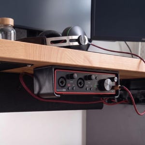 May include: A black Scarlett 2i2 audio interface mounted under a wooden desk. The device features a red trim, two input ports, gain control knobs, and a large volume dial. A red braided cable is plugged into the front, and black headphones rest on the desk above.