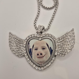 Puede incluir: Un collar con colgante en forma de corazón plateado que presenta una imagen central de una cara de cerdo. El marco del corazón y las alas adjuntas están incrustados con pequeños diamantes de imitación transparentes. El colgante cuelga de una cadena plateada.