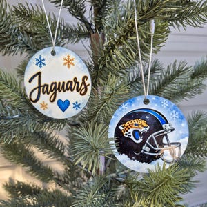 May include: Two round, flat Christmas ornaments hanging from silver strings. One ornament features the word Jaguars in black script with blue and orange snowflakes and a blue heart. The other displays a black football helmet with a jaguar logo.