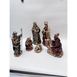 May include: A nativity set featuring six figurines including a shepherd with a staff, a kneeling figure, a baby in a manger, and three figures in robes, some adorned with small blue and red gems, finished in metallic gold, green, and red tones.