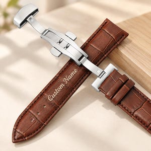 Könnte beinhalten: Ein braunes Leder-Uhrenarmband mit Krokodilprägung und einer silberfarbenen Schmetterlings-Faltschließe aus Edelstahl. Das Armband ist mit dem eingravierten Text Custom Name versehen und weist sichtbare Nähte sowie Einstelllöcher auf.