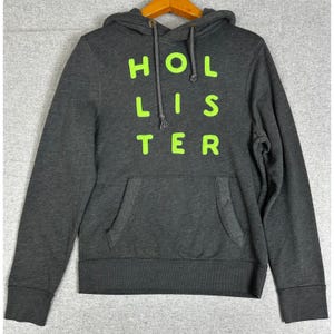 May include: A dark charcoal gray long-sleeve hooded sweatshirt featuring the text HOLLISTER printed in bright neon green block letters across the chest. The hoodie includes a front kangaroo pocket, a drawstring hood, and ribbed cuffs.