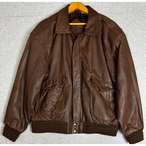 May include: A dark brown leather jacket featuring a classic collar, long sleeves with ribbed cuffs, and a ribbed hem. The jacket includes two large front pockets with flaps and a central zipper closure with two visible metal buttons at the bottom.