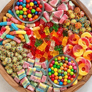 May include: A large wooden circular tray filled with an assortment of colourful gummy sweets, including gummy bears, watermelon slices, sour worms, rainbow squares, peach rings, and two glass bowls filled with small round multi-coloured sweets.