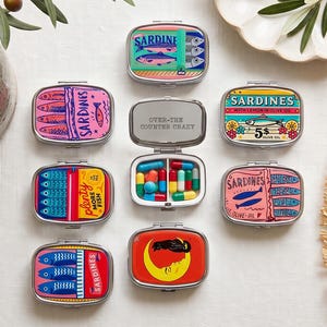 Pode incluir: Uma coleção de oito caixas de comprimidos retangulares de metal com tampas articuladas, apresentando designs gráficos coloridos com tema de sardinha. Uma caixa está aberta, revelando um compartimento interno branco cheio de várias cápsulas.