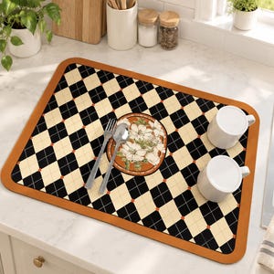 May include: A rectangular kitchen drying mat featuring a black and cream diamond argyle pattern with small orange accents and a solid orange border. In the center, a circular graphic displays white flowers. Two white mugs, a fork, and a spoon are placed on top.