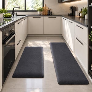 May include: Two rectangular, dark gray kitchen floor mats with rounded corners. The mats feature a solid, textured surface and are positioned on the floor in a kitchen with white cabinets, a black oven, and a black countertop.