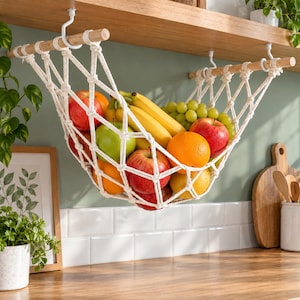 May include: A cream-colored macrame fruit hammock suspended from two wooden rods by white hooks. The net holds a variety of fresh produce, including red apples, green apples, yellow bananas, green grapes, and oranges, creating a hanging storage display.