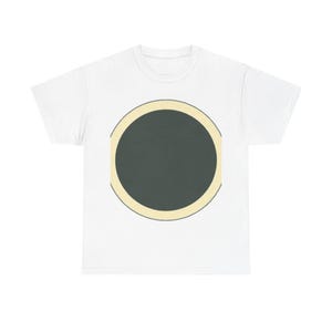 May include: A white short-sleeved t-shirt featuring a large, centred graphic design. The graphic consists of a solid dark charcoal grey circle enclosed within a thick, pale cream-coloured circular border, creating a bold, geometric focal point.