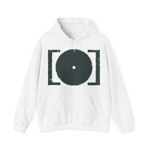 Op de afbeelding: Een witte hoodie met een grafische print op de borst. Het ontwerp bestaat uit een grote, donker antracietgrijze cirkel gecentreerd tussen twee verticale, getextureerde antracietgrijze haakjes, met een kleine, lichtgele stip in het midden.