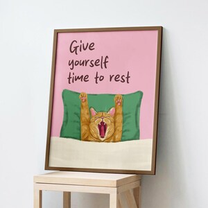 Puede incluir: Una lámina enmarcada con fondo rosa que muestra a un gato naranja bostezando con las patas levantadas, tumbado sobre una almohada verde bajo una manta blanca. El texto dice, Give yourself time to rest, en letras manuscritas marrón oscuro.