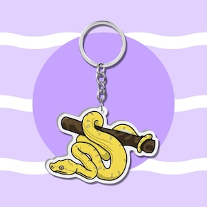 May include: A keyring featuring a yellow snake wrapped around a brown branch. The snake has a light yellow underbelly and darker yellow scales. The charm is attached to a silver metal split ring by a short silver chain, creating a functional accessory.