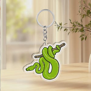 May include: A clear acrylic keychain featuring a vibrant green snake coiled around a brown wooden branch. The snake has a smooth, bright green body with a darker green head and black eye detail. The keychain includes a silver metal ring and chain.